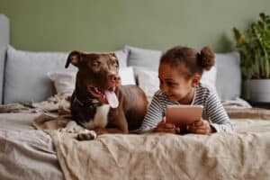 Happy dog and young smiling individual with a tablet hanging out on a bed together
