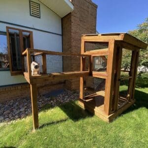 Wooden box-like structure that is screened in with a tunnel of the same materials out a window of a home to create a catio cat enclosure